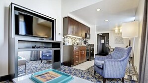 A modern hotel room with a flat-screen TV, a kitchen area with wooden cabinets, a dining table with a tray, and a blue patterned carpet.