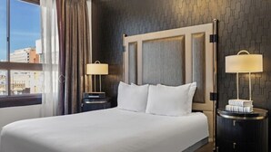 A hotel room with a large bed, two bedside tables with lamps, a view of the city through a window with curtains, and a textured wall.