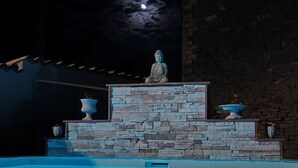 A nighttime pool area with a statue on a stone platform, surrounded by stone walls and a clear pool.