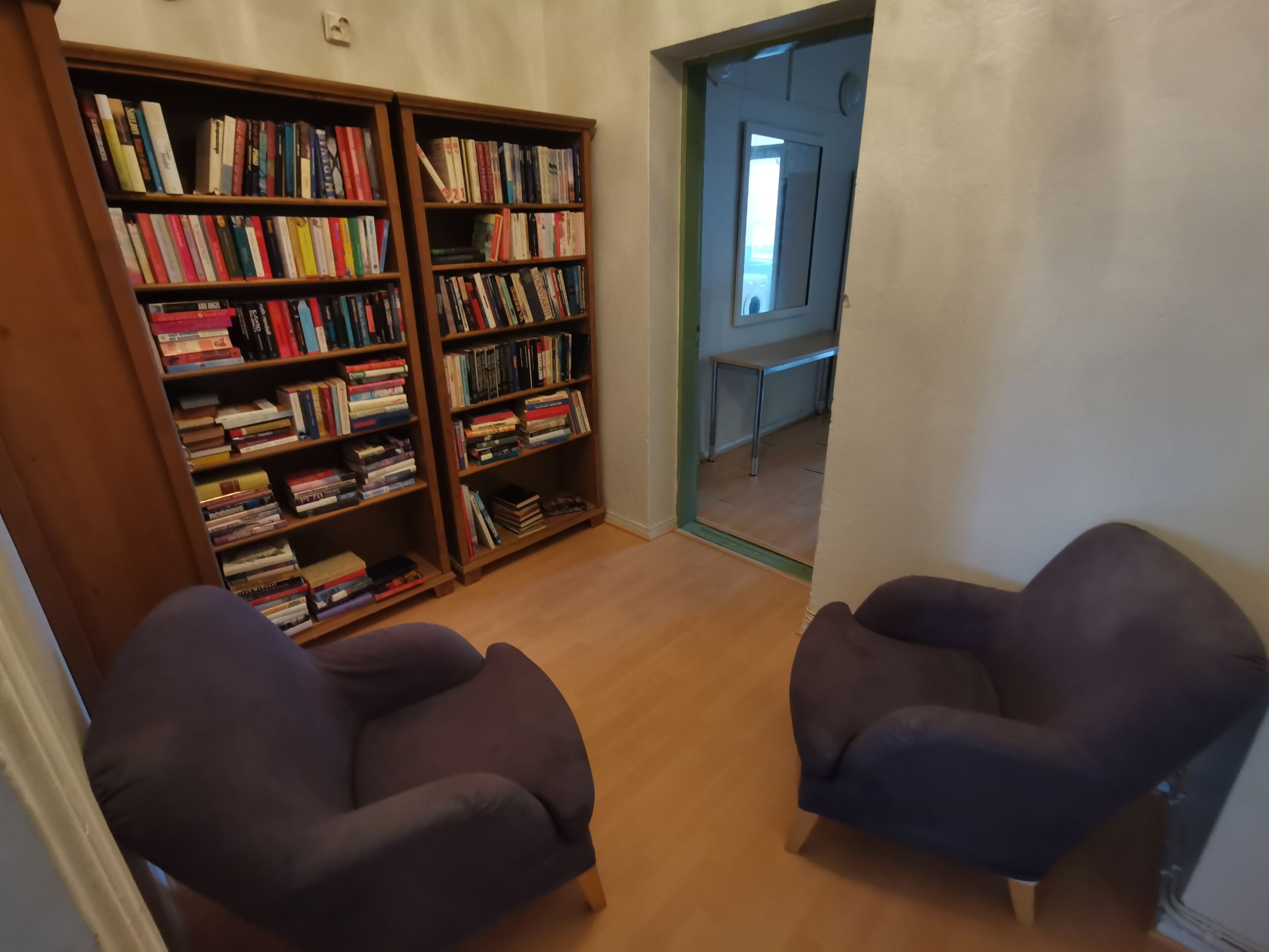 A room with two dark armchairs, a wooden bookshelf filled with books, and a view of a hallway with a bench.