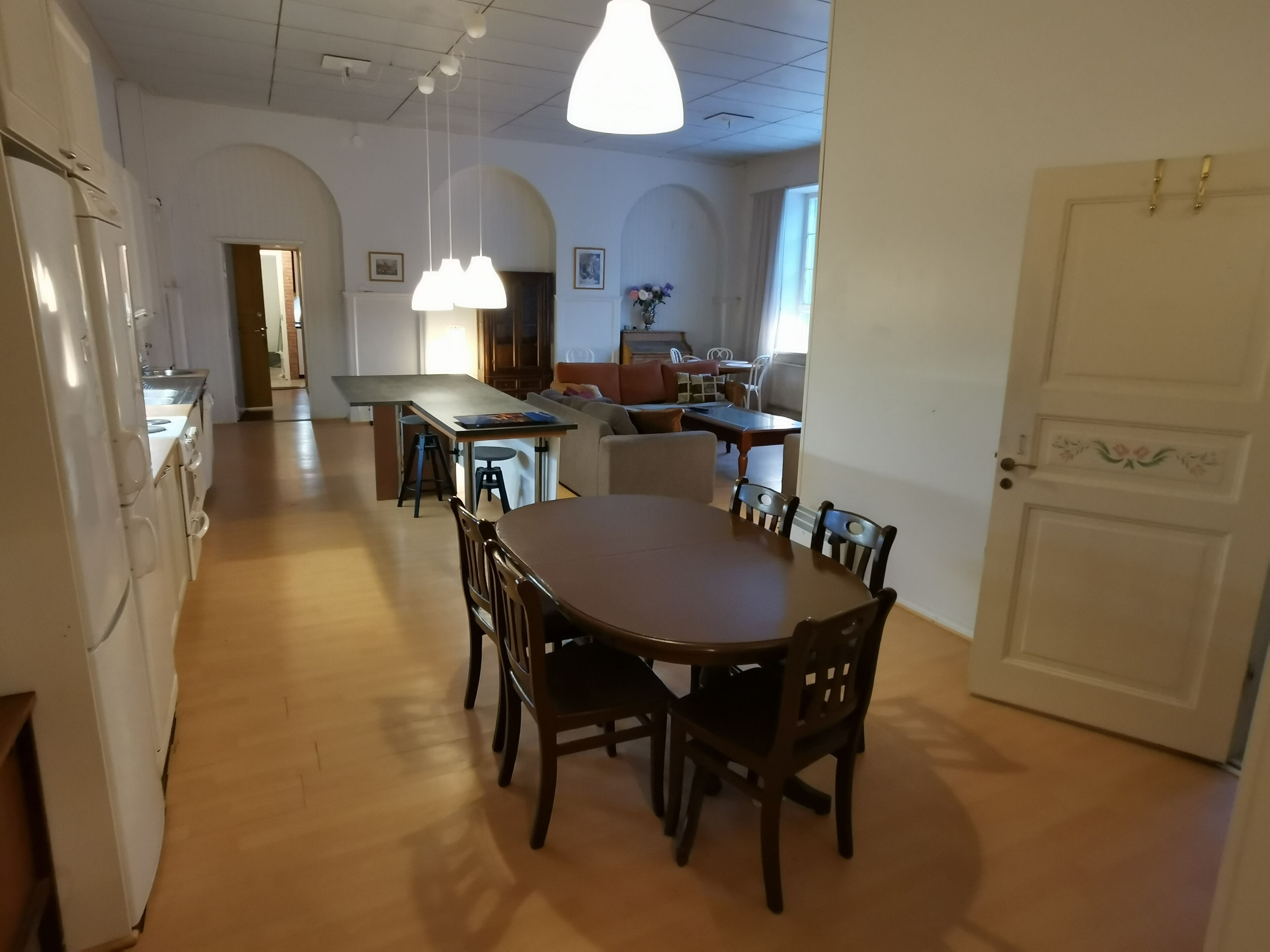Un coin repas avec une table et des chaises en bois, une cuisine en arrière-plan et une porte blanche dotée d’une poignée décorative.