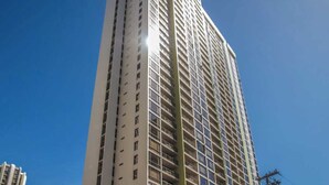 A tall, modern building with multiple floors and balconies, surrounded by other buildings and a clear sky.