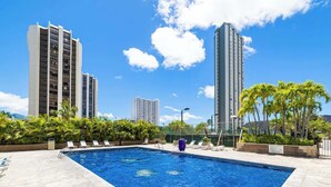A swimming pool with lounge chairs, surrounded by palm trees and tall buildings.