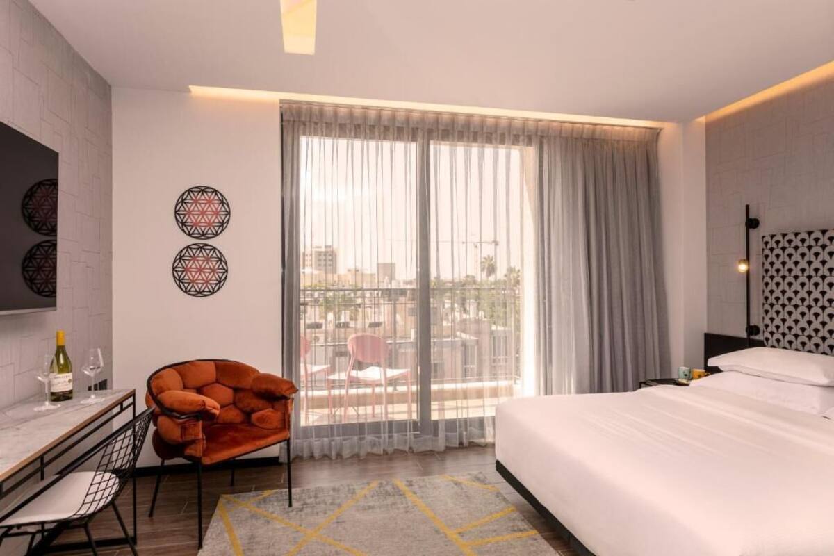 A hotel room with a bed, a desk with a chair, a bottle of wine, and a view of the city through large windows.