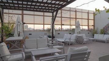 An outdoor patio with white furniture, including sofas and tables, under a pergola with a view of the cityscape.