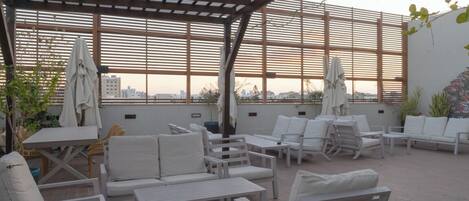 An outdoor patio with white furniture, including sofas and tables, under a pergola with a view of the cityscape.