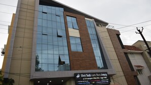 Un edificio moderno con un letrero que indica Residencia Shri Subramaniam y Salón Mini Ishwaram para Bodas, que presenta una amplia fachada de vidrio y una combinación de tonos beige y marrón.
