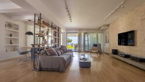 A modern living room with a sofa, coffee table, and TV against a brick wall. There are shelves with bottles and decorative items, and a dining area with chairs and a table. - Spacious beach front Vila with 100 sqm infinity pool,  a basket ball court (Rhodes)