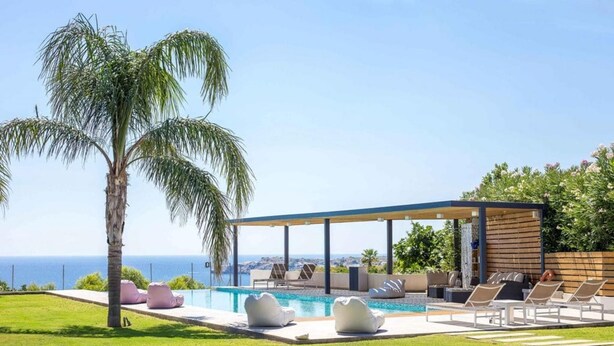 A modern outdoor pool area with lounge chairs and a covered seating area, overlooking a clear blue sky and a distant view of the sea. - Spacious beach front Vila with 100 sqm infinity pool,  a basket ball court (Rhodes)