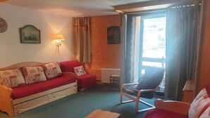 A cozy living room with a wooden floor, a red sofa, a wooden coffee table, and a chair. There is a window with curtains and a radiator on the wall. - Charming 32m² studio, balcony, near slopes, Les Menuires (Les Menuires)