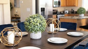 Een eettafel gedekt met witte borden, een potplant en kandelaars in een moderne keuken met houten kasten en een roestvrijstalen koelkast.