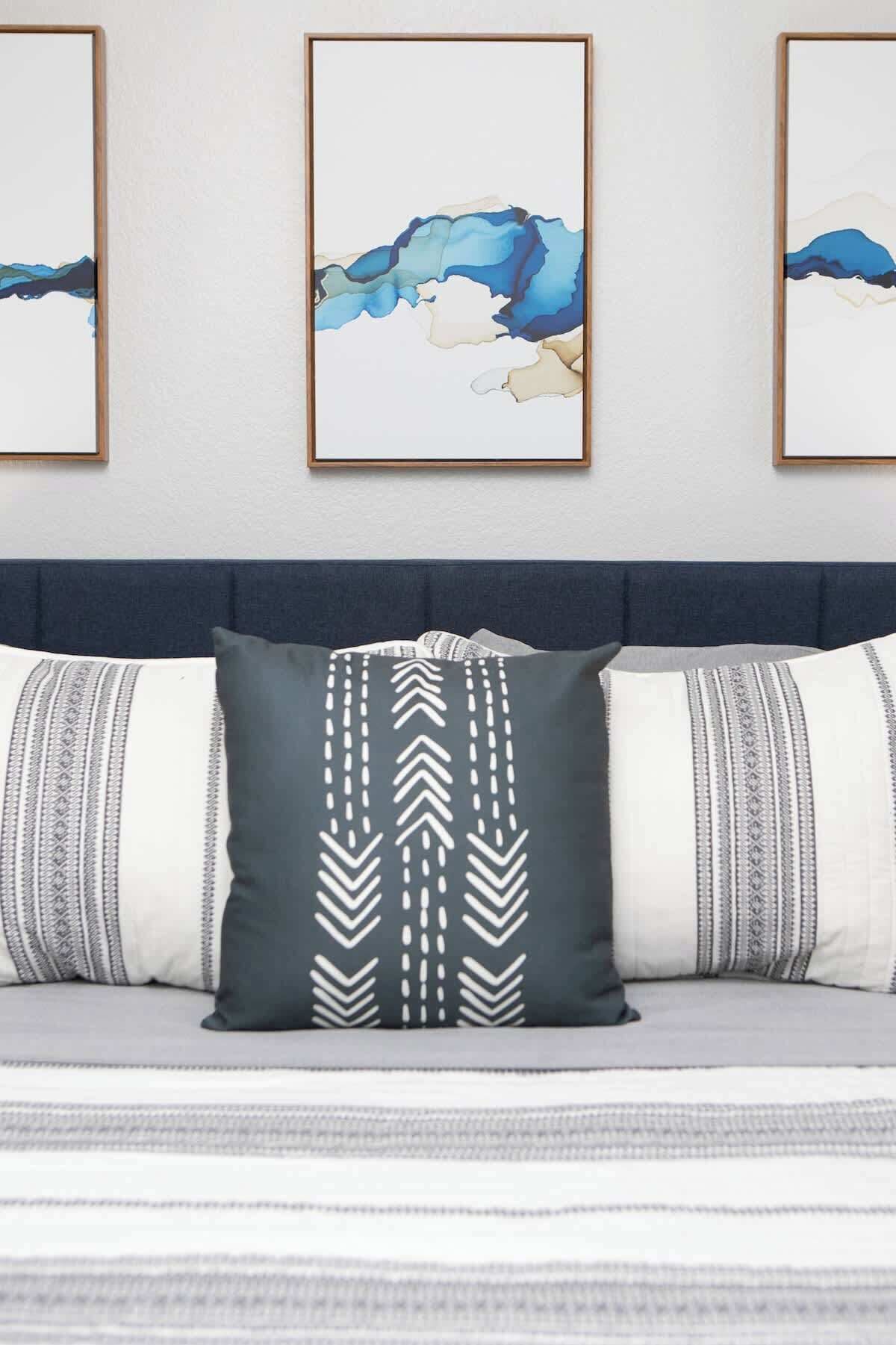 A bedroom with a bed, a dark grey pillow with a white chevron pattern, and two framed abstract paintings on the wall.