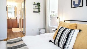 A modern bedroom with a bed, a bedside table, a window with curtains, a plant on a shelf, and a view of the outside. - Royal Skyline -Spacious & Modern 3BR w Pool (Henderson)