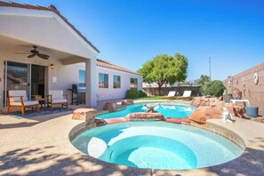 Une piscine dont le centre est plus profond que le reste, entourée de rochers et dotée d’un patio couvert avec des chaises et un ventilateur de plafond.