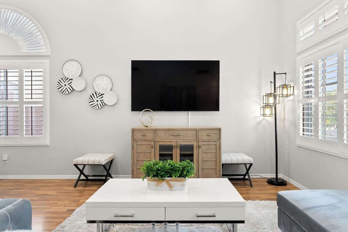 A modern living room with a flat-screen TV mounted on the wall, a wooden cabinet with drawers, two cushioned stools, a white coffee table with a plant, and a floor lamp.