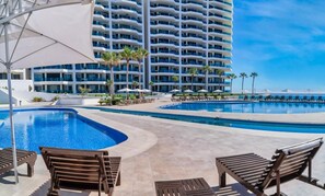 Ein Hotelpoolbereich mit mehreren Schwimmbecken, Liegestühlen und einem großen Gebäude im Hintergrund.