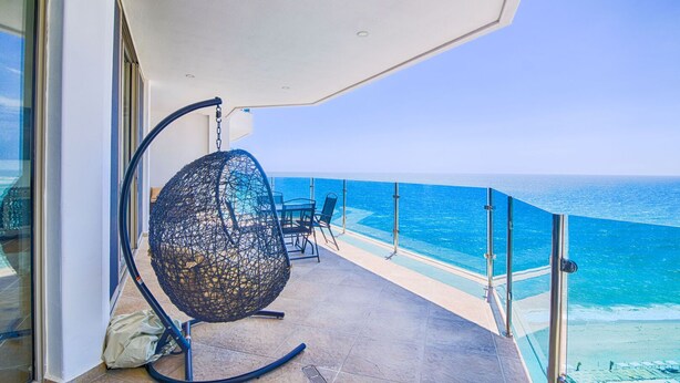 Un balcón con una hamaca colgante, una mesa y sillas, con vista a un mar azul y despejado.