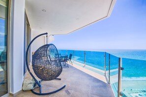 A balcony with a hanging chair, a table, and chairs, overlooking a clear blue sea.