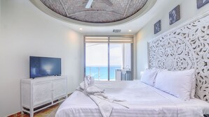Habitación de hotel con una cama grande, un televisor sobre un mueble blanco y vista al mar a través de una puerta corrediza.