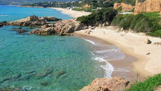 A coastal landscape with clear waters, a sandy beach, rocky outcrops, and distant hills.