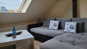 A loft-style room with a sloped ceiling, a grey sofa with decorative pillows, a small table with a blue bottle, and a view of the sky through a skylight. - Vacation apartment RuBi - Holiday apartment (Stralsund)