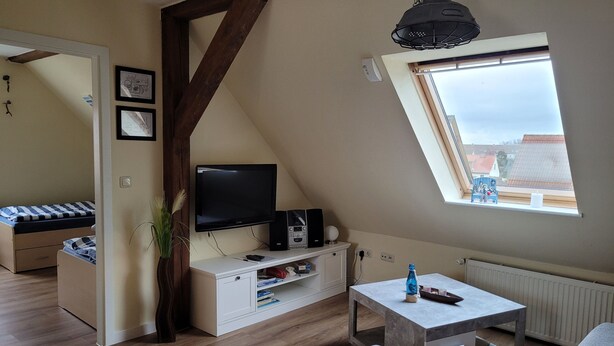 A loft-style room with a sloped ceiling, a television, a white cabinet, a small table, and a window with a view of buildings. - Vacation apartment RuBi - Holiday apartment (Stralsund)