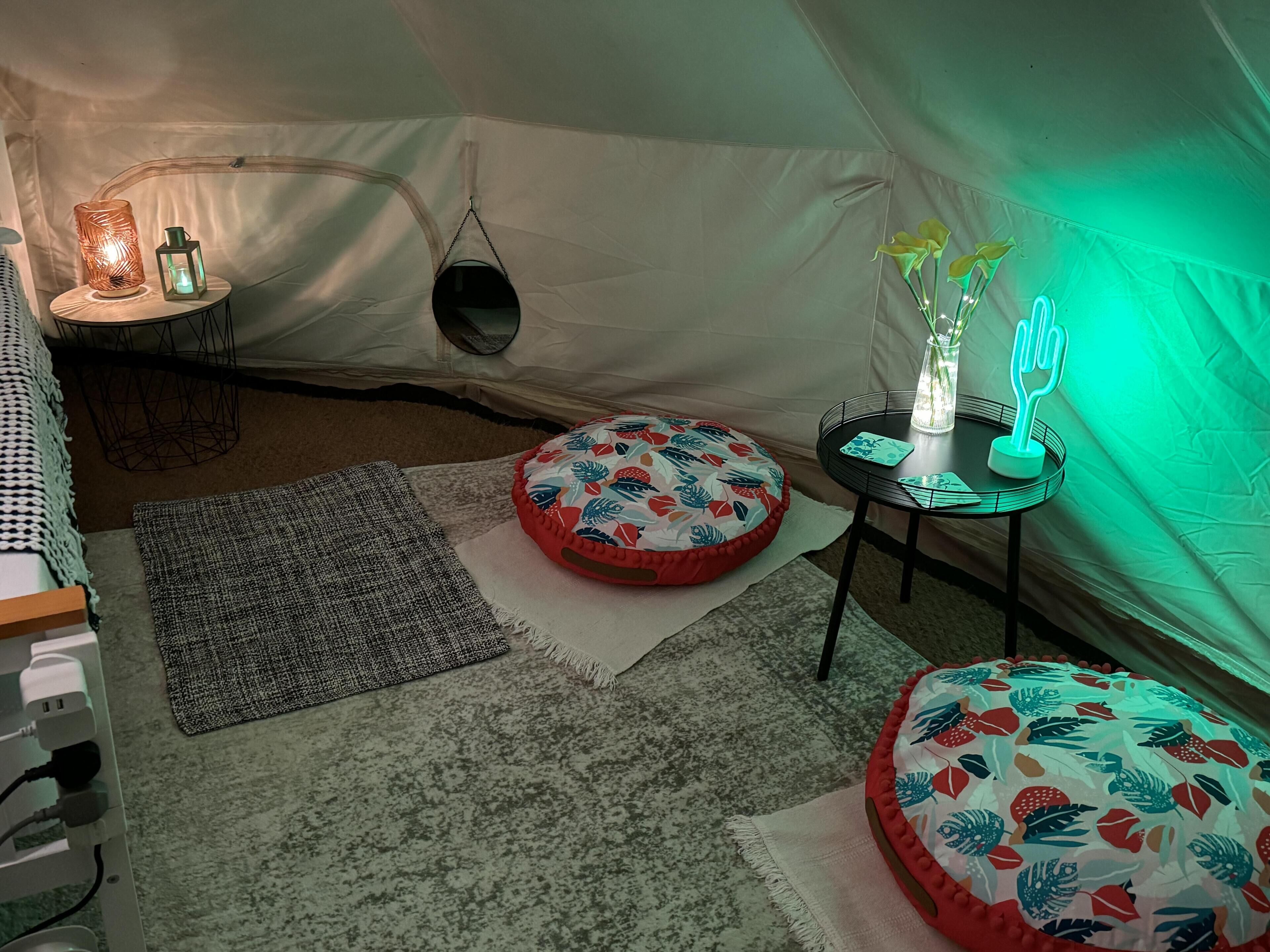 A cozy tent interior with a round table, two ottomans, a hanging chair, and a small lamp.