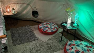 A cozy tent interior with a round table, two ottomans, a hanging chair, and a small lamp.