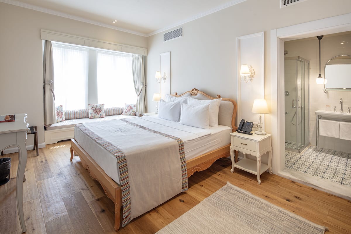 Hotellihuone, jossa on suuri sänky, yöpöytä lampun kanssa, suihkutila ja ikkuna, jossa on verhot.