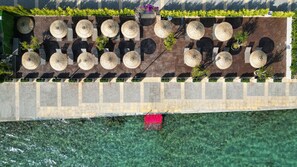 An aerial view of a waterfront patio with multiple round tables and chairs, a red barrier, and clear water.