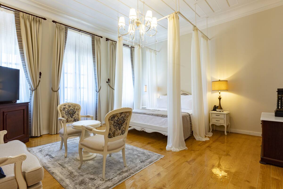 Hotellihuone, jossa on sänky, televisio, ruokapöytä tuoleineen, kattokruunu ja verhot.