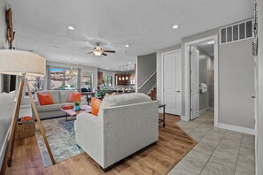 A modern living room with a sofa, coffee table, and a lamp. There is a staircase leading to a kitchen area with cabinets and a dining table.