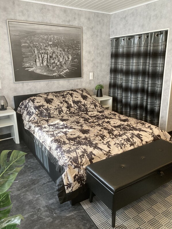 A bedroom with a bed, a nightstand, a potted plant, a large window with curtains, and a wall-mounted picture.