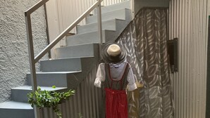 Una escalera con barandilla de metal, plantas en macetas y un maniquí vestido con un delantal rojo y una blusa blanca.