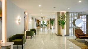 Een moderne hotellobby met een marmeren vloer, groene fauteuils, een receptiebalie en potplanten.