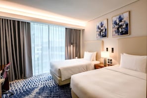 A hotel room with two beds, a desk, a lamp, and a large window with curtains.