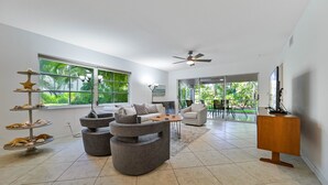 Una sala de estar moderna con un sofá seccional gris, una mesa de centro, un mueble para televisión y una exhibición de estrella de mar. Amplias ventanas ofrecen vista a zonas verdes exteriores.