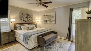 A bedroom with a bed, bedside tables, a bench, a ceiling fan, artwork, and a window with curtains.
