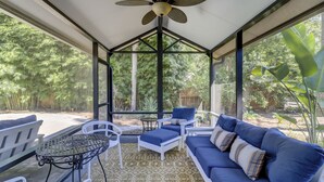 A covered outdoor seating area with a ceiling fan, white and blue furniture, and a view of a garden.