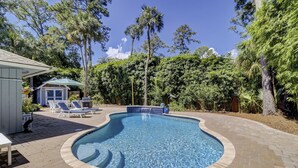 A swimming pool with a curved edge, surrounded by a brick patio. There are lounge chairs and a small building with a blue roof.