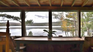 A wooden table with a potted plant, a chair, and a view of a lake and trees through large windows. - "Andy's Place" (Abbot)