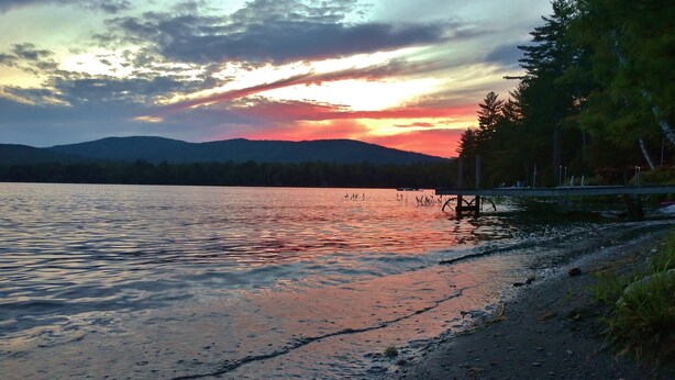 A lakeside view with a sunset, calm water, and a forested shoreline. - "Andy's Place" (Abbot)