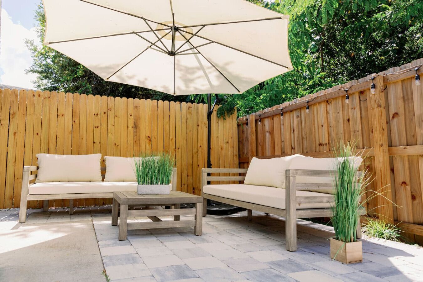 Un'area patio esterna con recinto in legno, due divani beige, un tavolo in legno e un ombrellone.
