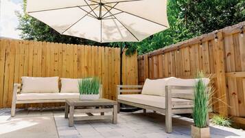 Un'area patio esterna con recinto in legno, due divani beige, un tavolo in legno e un ombrellone.