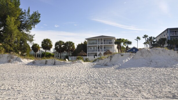 A beachfront property with a large building featuring multiple balconies, a sandy beach in the foreground, and palm trees in the background.