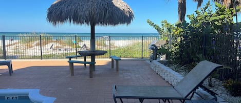 Zona junto a la piscina con una sombrilla de paja, sillas de descanso y vista a la playa.