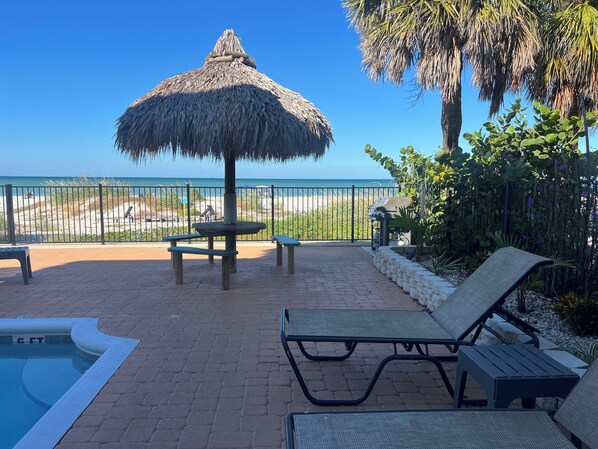 Zona junto a la piscina con una sombrilla de paja, sillas de descanso y vista a la playa.