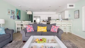 A living room with a grey sofa, colorful cushions, a coffee table with fruits, and a kitchen in the background.