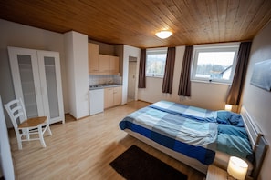 A bedroom with a bed, a chair, a wardrobe, a small kitchenette, and a window with curtains.