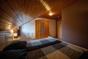 Een slaapkamer met een houten plafond, een bed met een geruit dekbed, een houten garderobe en een nachtkastje met een lamp.